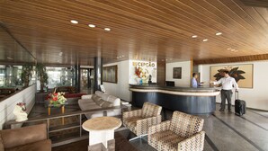 A hotel reception area with a wooden ceiling, a reception desk, a sofa, patterned chairs, and a reception counter. - Xenius Hotel (João Pessoa)