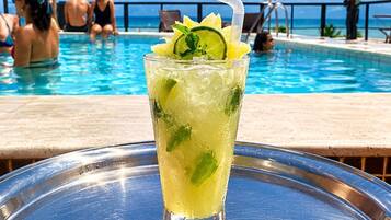 Un bicchiere di bevanda tropicale con lime e menta su un vassoio, con una piscina e persone sullo sfondo.