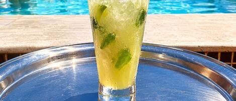 A glass of a tropical beverage with lime and mint on a tray, with a pool and people in the background.