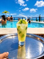 Un bicchiere di bevanda tropicale con lime e menta su un vassoio, con una piscina e persone sullo sfondo.