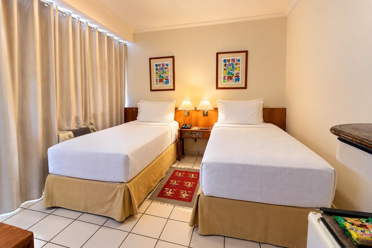 Habitación de hotel con dos camas individuales, una mesita de noche con lámpara, una pequeña mesa con silla y una ventana con cortinas.