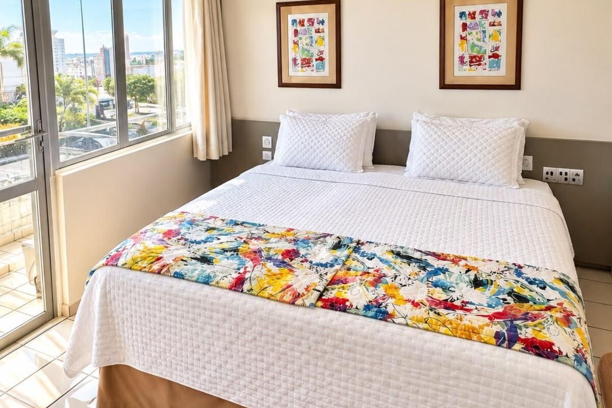 A hotel room with a large bed, a window with a view of the city, and two framed artworks on the wall.
