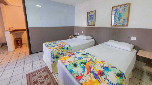 A hotel room with two beds, a small table, and a chair. There is a window and a painting on the wall. - Xenius Hotel (João Pessoa)