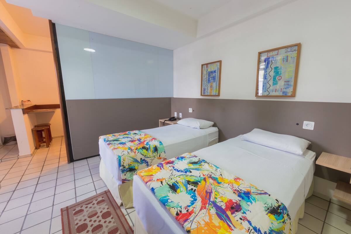 A hotel room with two beds, a small table, and a chair. There is a window and a painting on the wall.