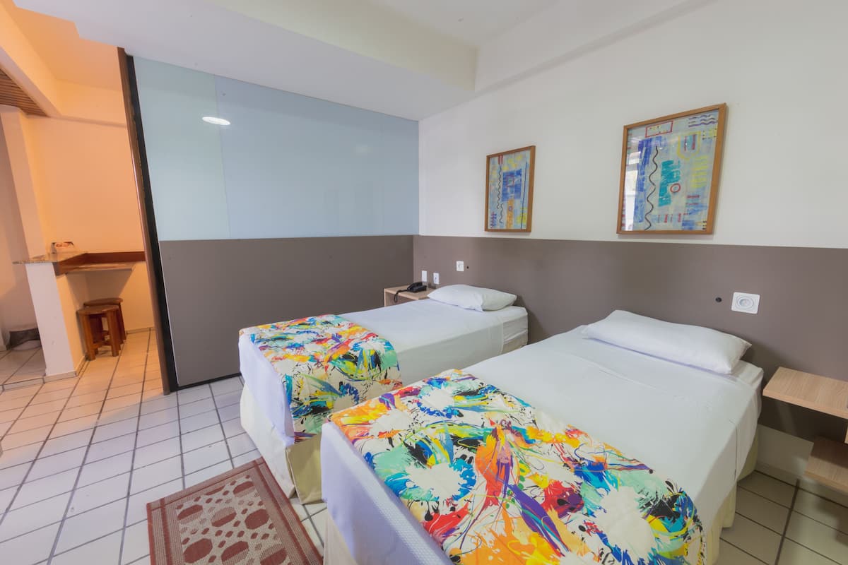 A hotel room with two beds, a small table, and a chair. There is a window and a painting on the wall.