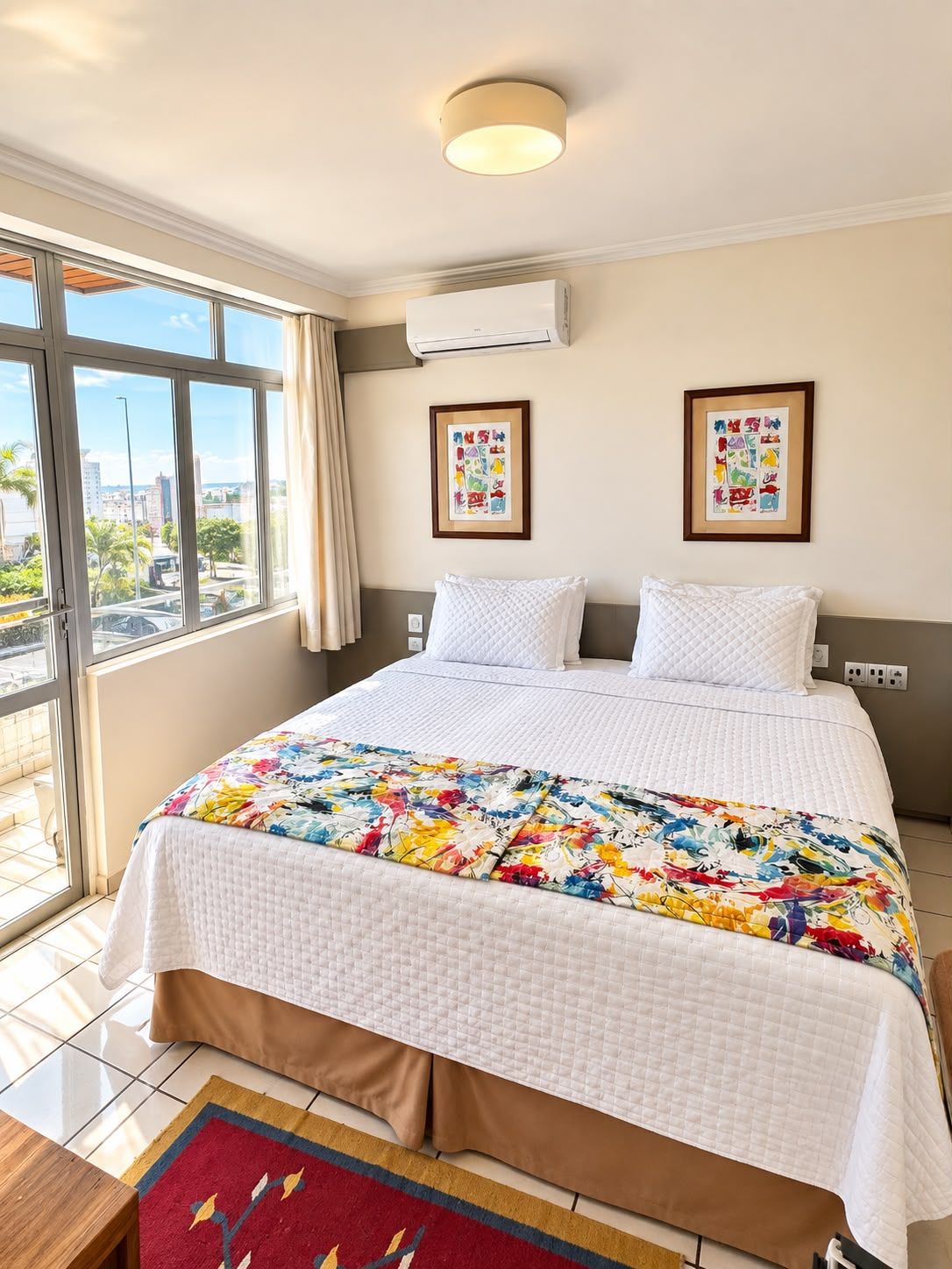 A hotel room with a large bed, two pillows, a colorful quilt, a window with a view of the city, and an air conditioning unit.
