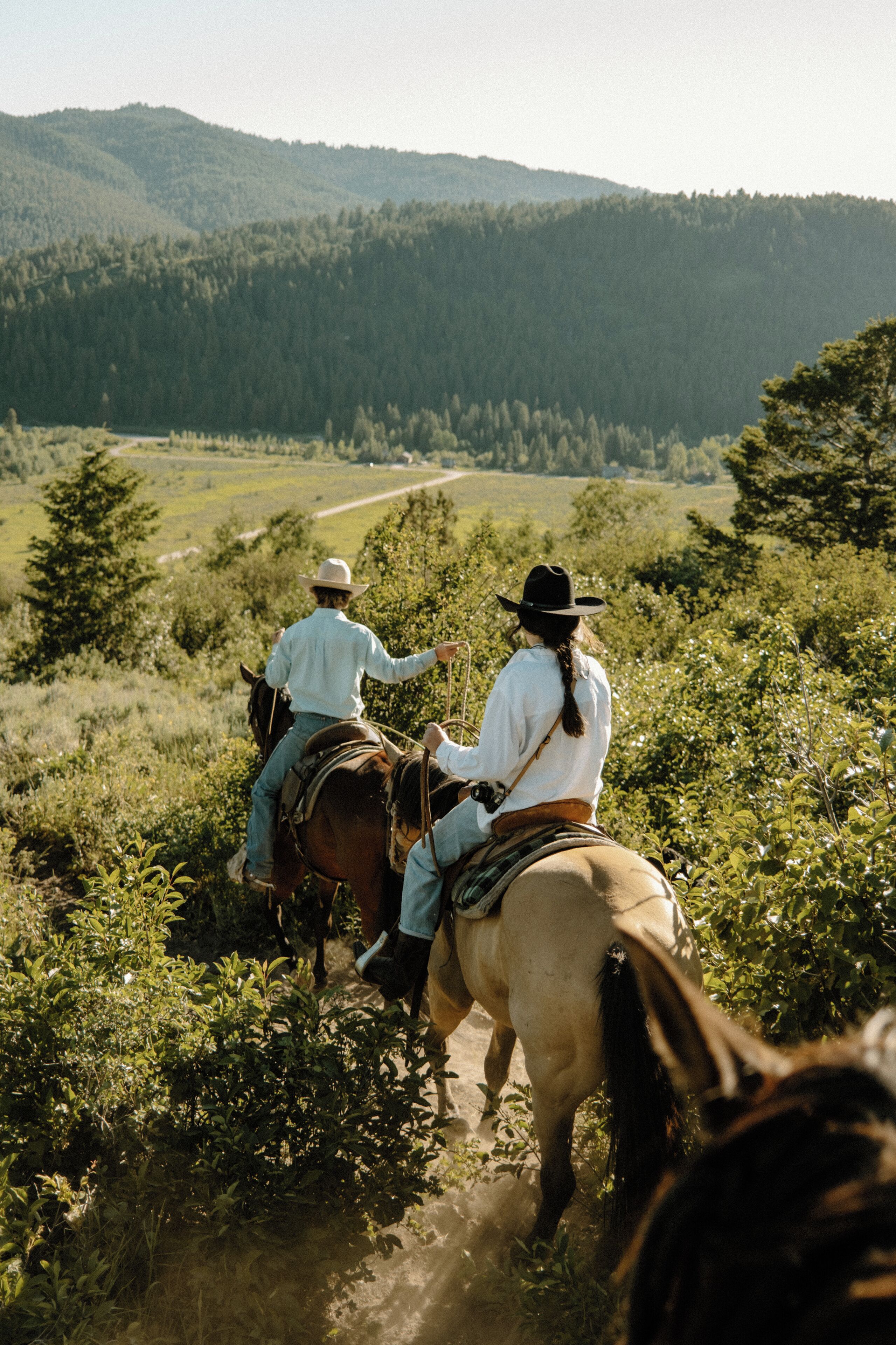 Dos personas cabalgando por un sendero de tierra rodeado de vegetación y colinas.