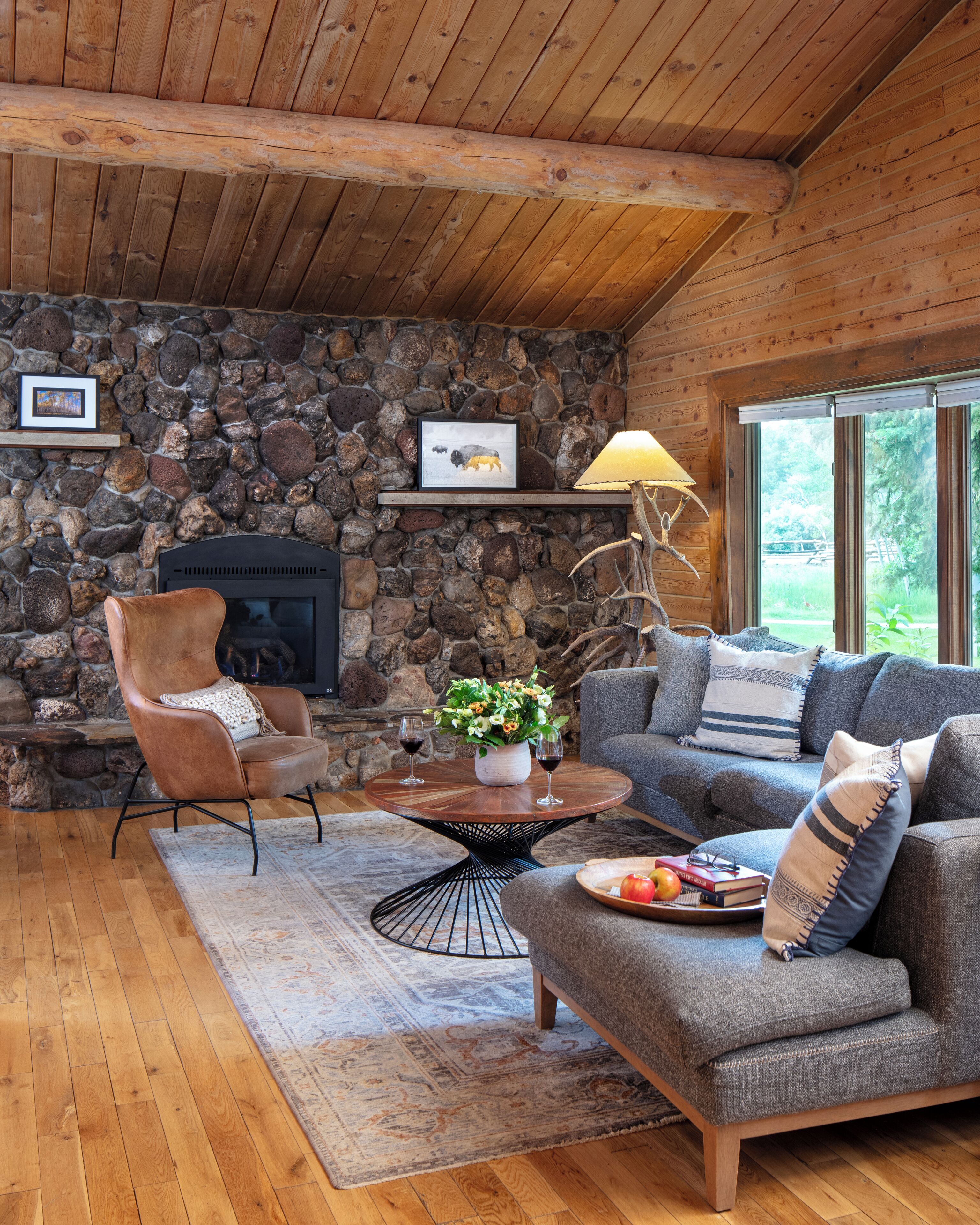 A cozy living room with a stone wall, a fireplace, a leather armchair, a grey sofa, and a coffee table with a vase of flowers.