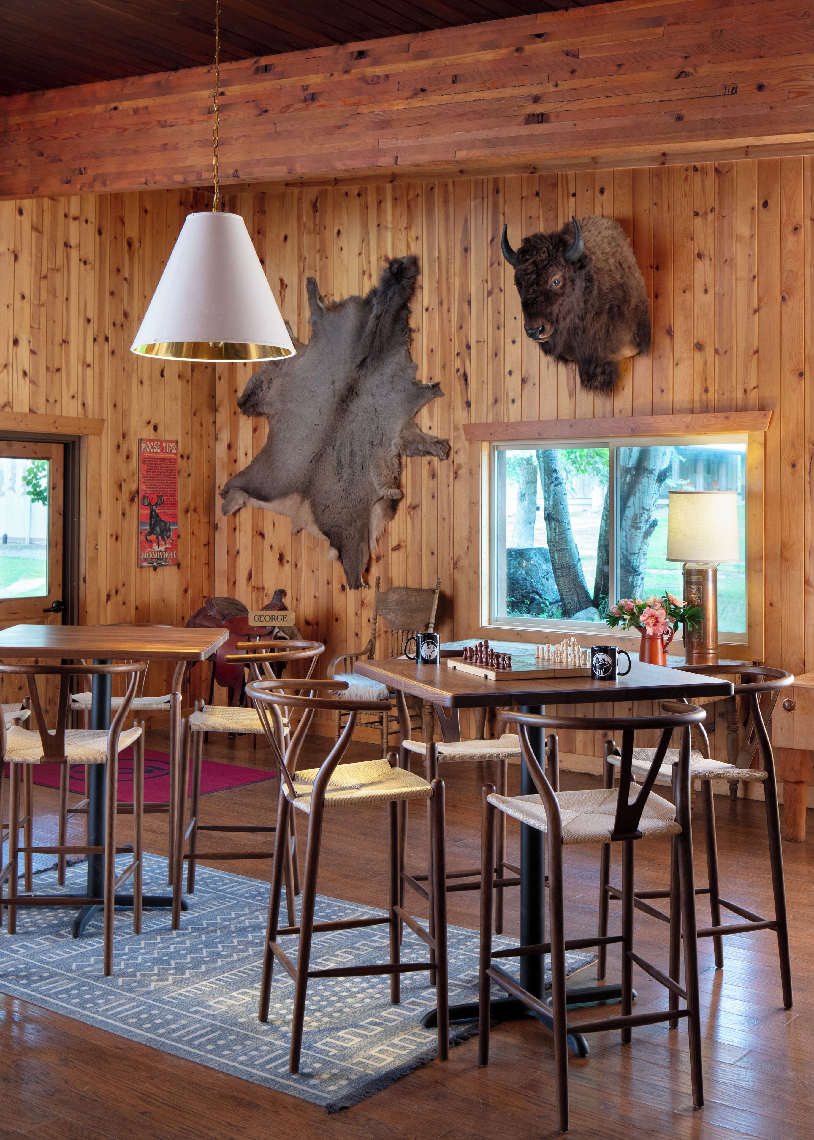 A rustic interior with wooden walls, a dining area with metal chairs, a table with chess pieces, and mounted animal heads.