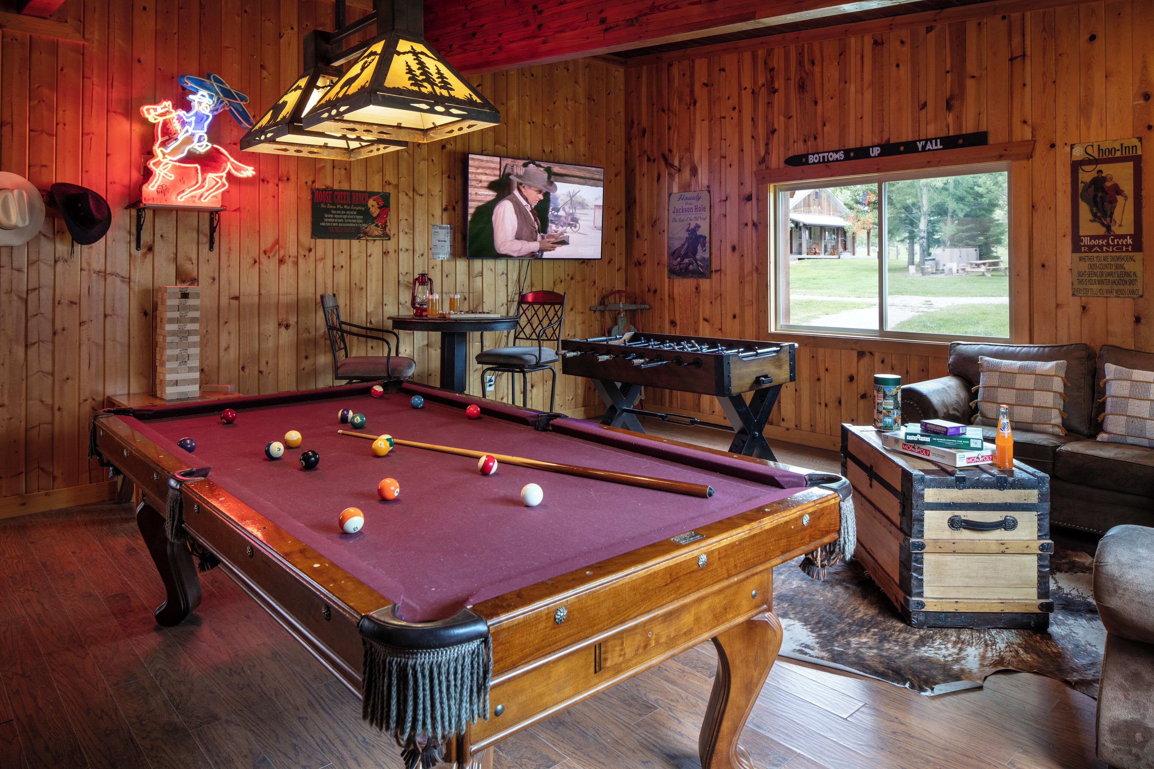 Une table de billard avec des billes, un néon, un téléviseur et une fenêtre donnant sur l’extérieur.