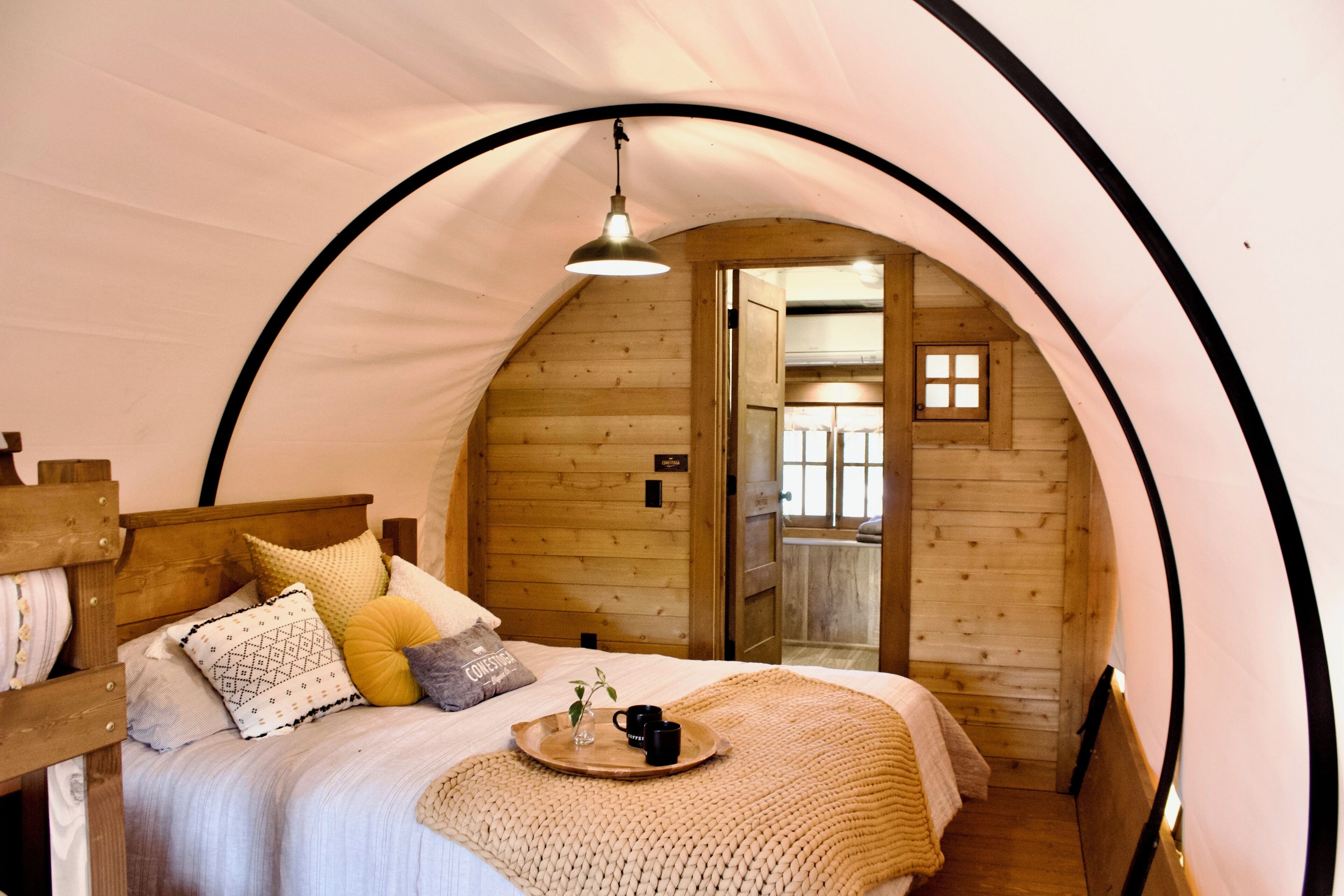 A cozy, circular tent-style room with a wooden bed, a tray with a mug and a plant, and a hanging light fixture.