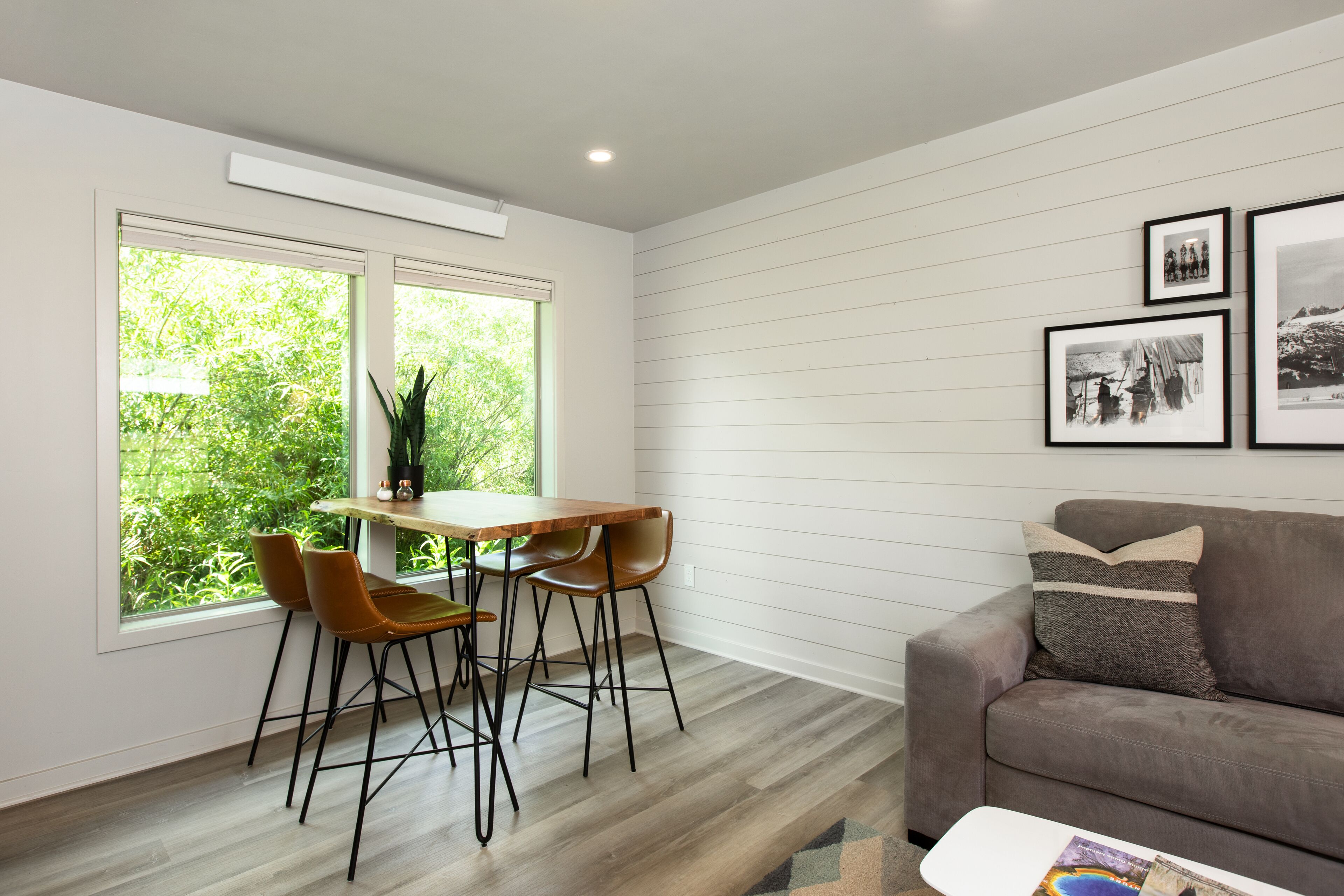 Ein modernes Wohnzimmer mit einem Holzesstisch, zwei Stühlen, einem Sofa und einem Couchtisch. Große Fenster bieten Blick auf Grünflächen außerhalb.