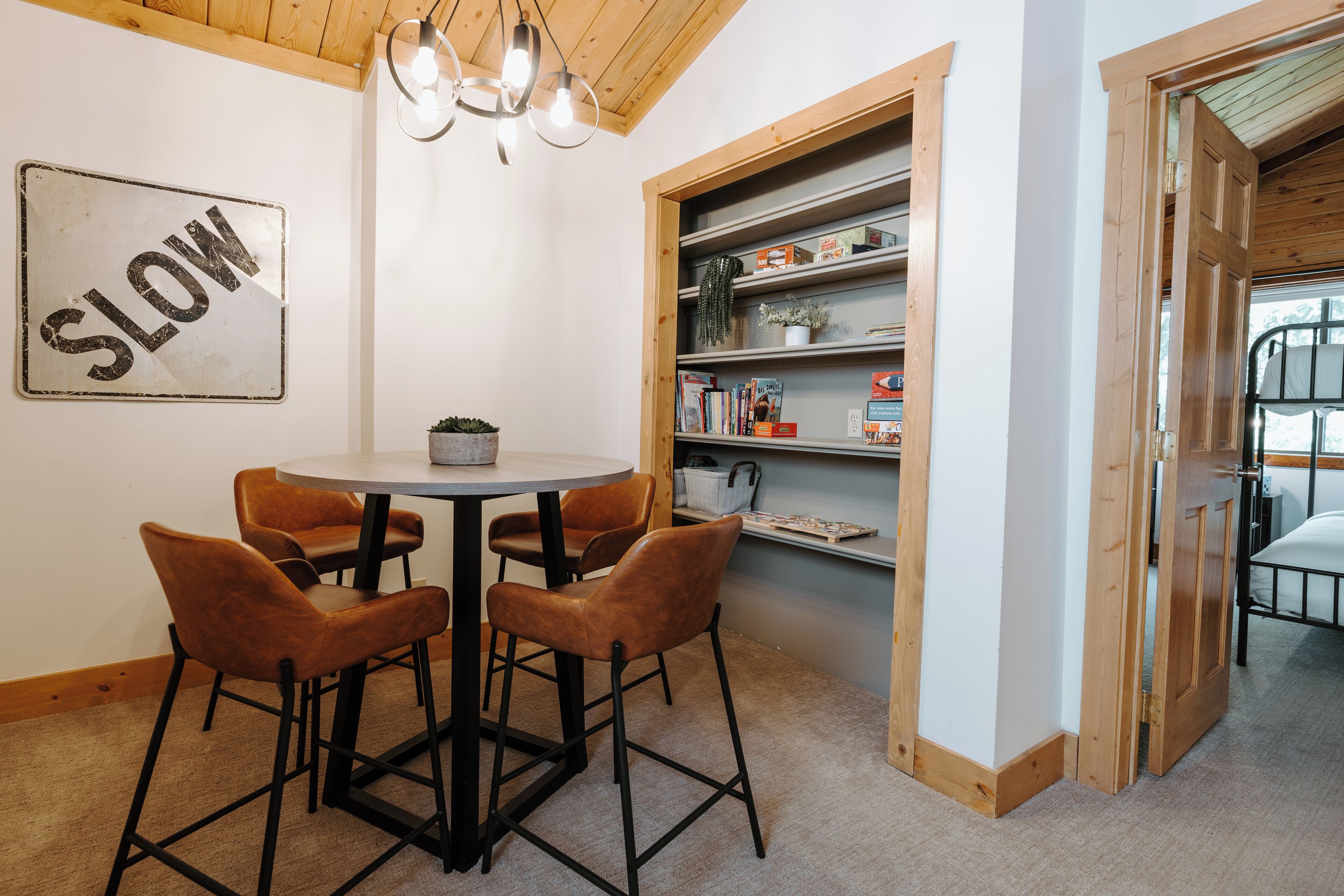 Una habitación acogedora con una mesa redonda y cuatro sillas, una estantería con libros y objetos decorativos, y un cartel que dice 'SLOW'.