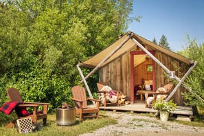 A rustic wooden cabin with a porch, two Adirondack chairs, a fire pit, and a checkered blanket.