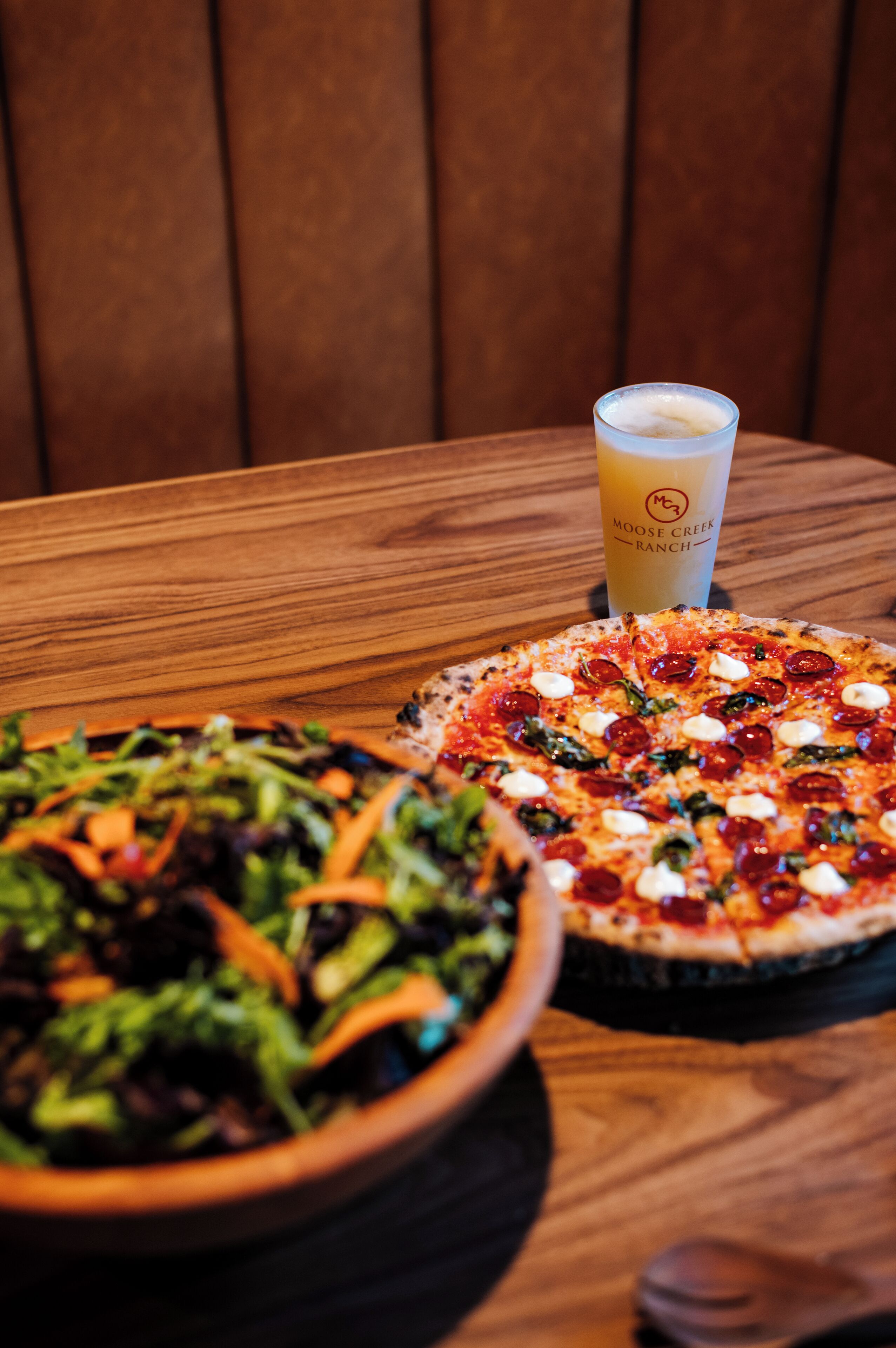 A pizza and a salad on a wooden table with a drink in the background.