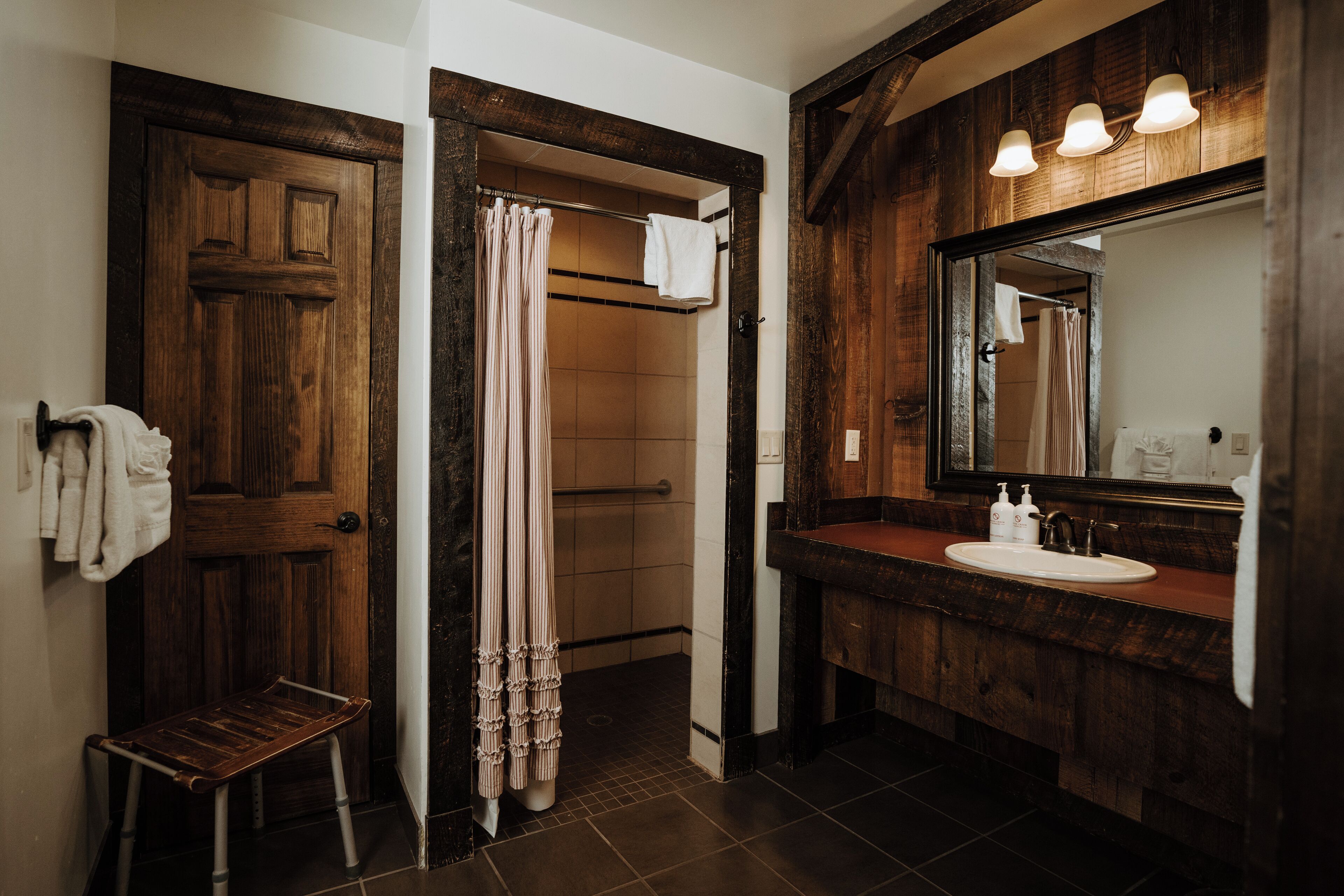 A bathroom with a wooden door, a shower with a curtain, a sink with a mirror, and a bench.