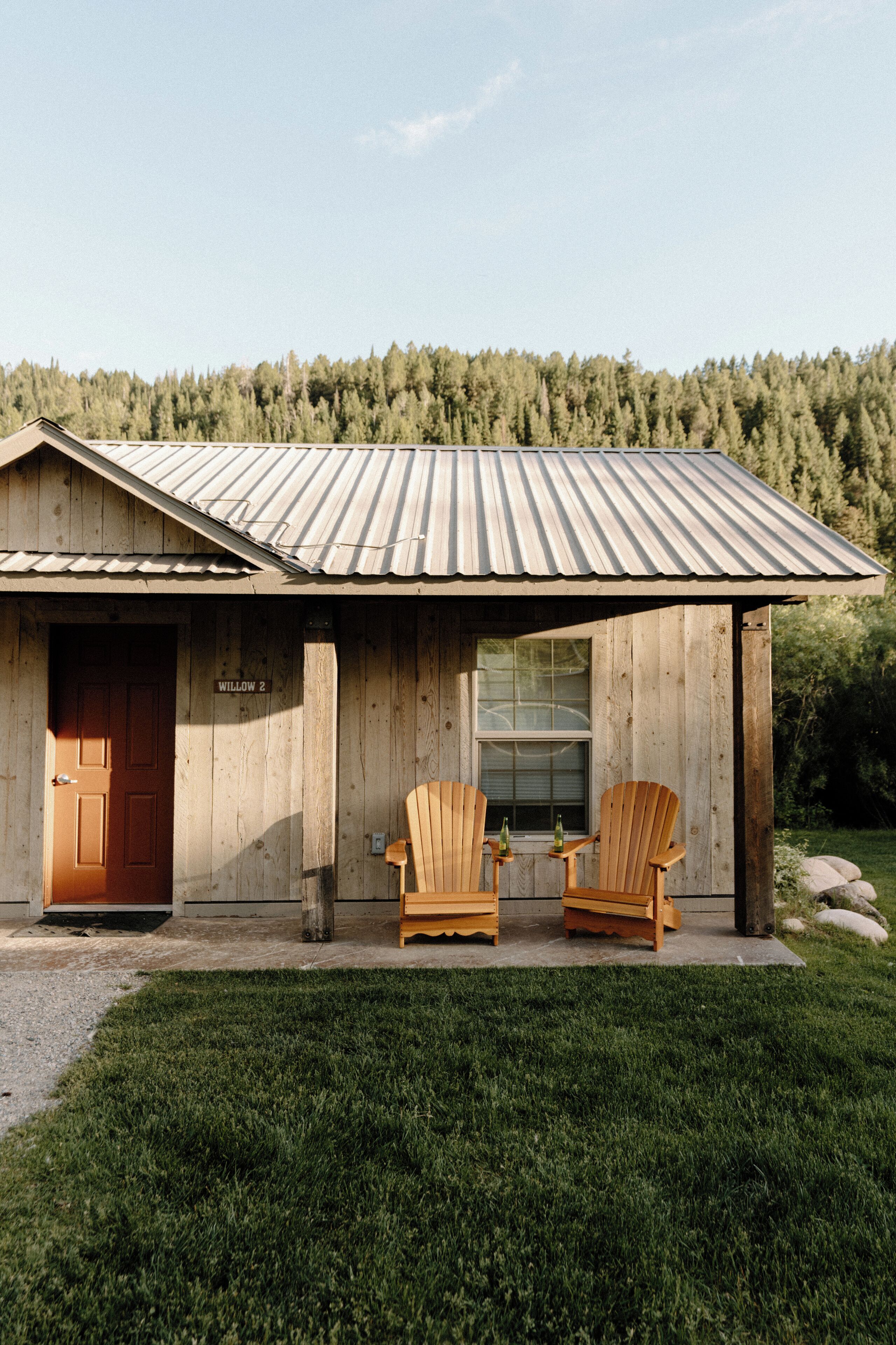 Une cabane en bois avec un toit en métal, deux chaises en bois, une petite table et une porte portant l’inscription « Saule 2 ».