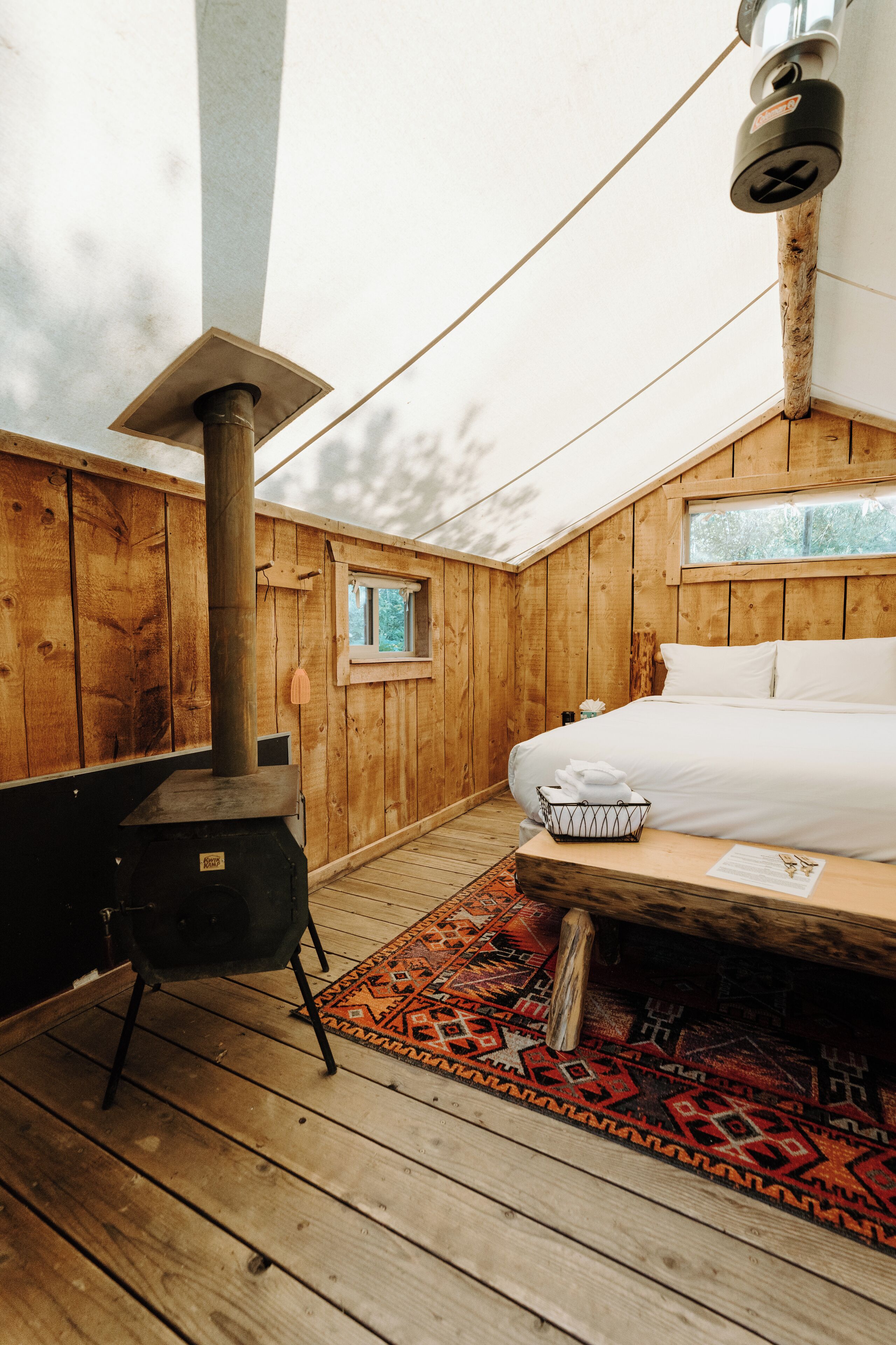 Ein Zimmer in einer Holzhütte mit einem Bett, einem kleinen Fenster, einem Heizgerät und einem gemusterten Teppich.