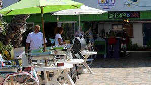 Un café en plein air avec des tables et des parasols, une boutique de souvenirs et une enseigne indiquant l’emplacement d’un magasin de surf.