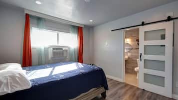 Un dormitorio con cama, una ventana con cortinas, un equipo de aire acondicionado y una puerta corrediza que da a un baño.