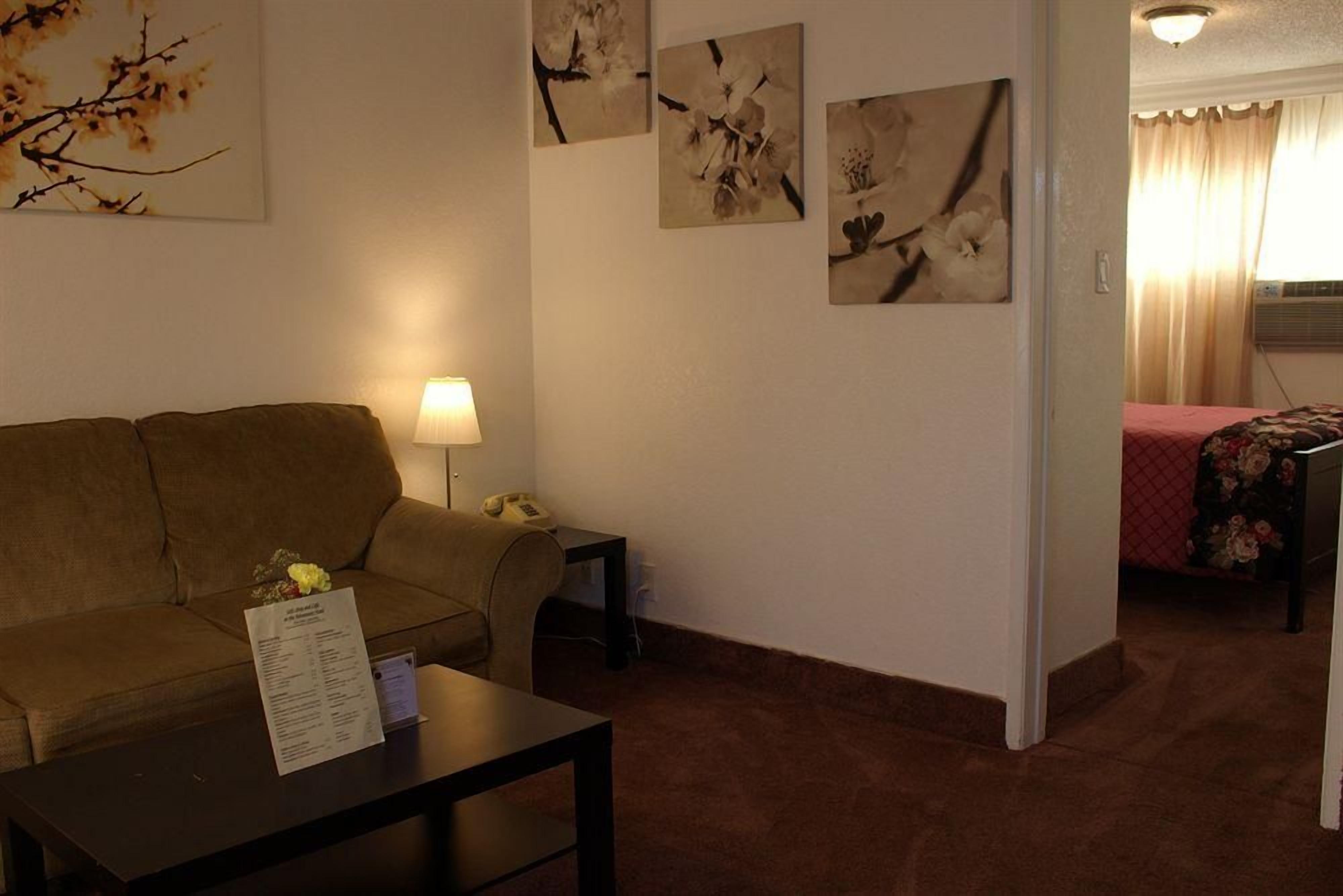 A room with a sofa, a small table with a lamp, and a bed with a floral bedspread. There are wall-mounted pictures of flowers.