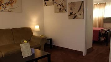 Una habitación con un sofá, una mesita de noche con una lámpara y una cama con colcha estampada con motivos florales. Hay cuadros de flores colgados en la pared.