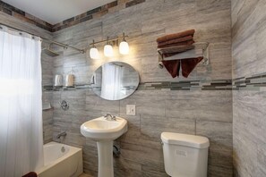 A bathroom with a bathtub, sink, and toilet, featuring a stone wall and a towel rack. - Tradewinds All Suite Hotel (Inglewood)