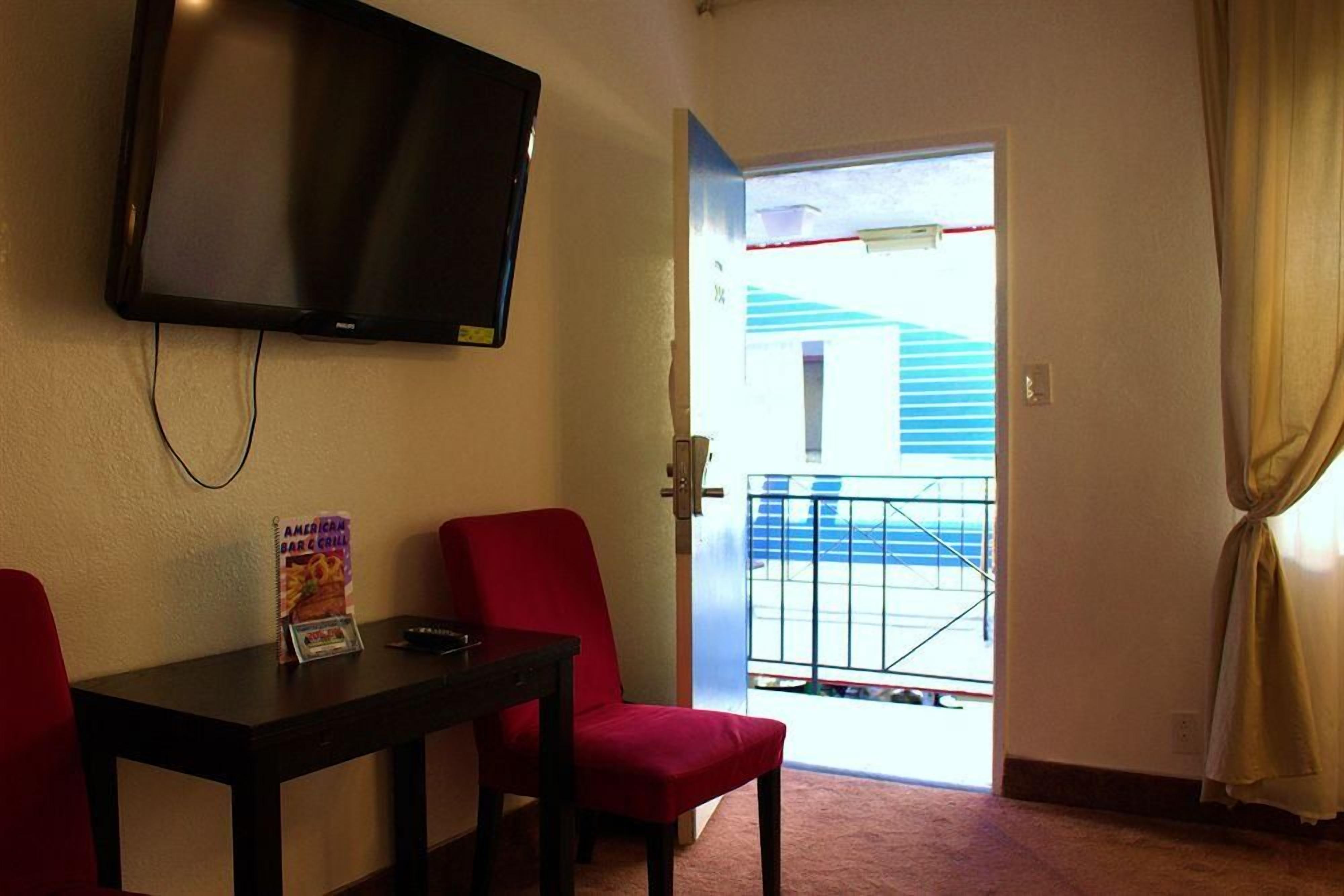 A room with a television mounted on the wall, a table with a chair, and a view of a balcony with a railing.