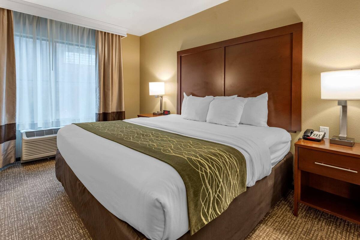 A hotel room with a large bed, a wooden headboard, a bedside table with a lamp, and an air conditioning unit.