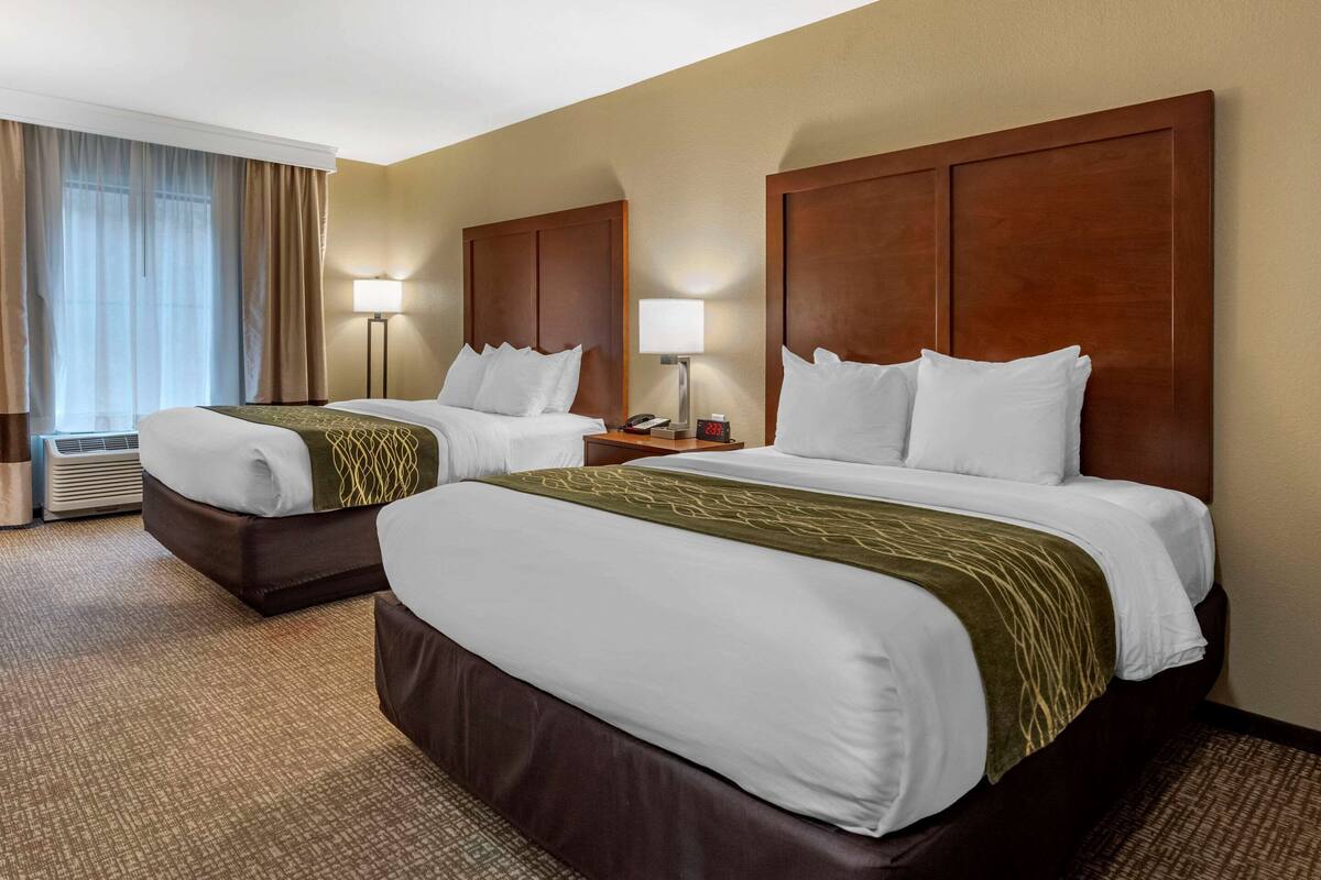 A hotel room with two beds, each with a wooden headboard, white bedding, and a green throw. There is a bedside table with a lamp and a clock, and a window with curtains.