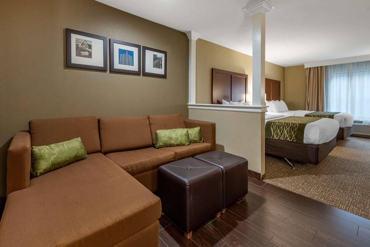A hotel room with a brown sectional sofa, two ottomans, a bed with a green and white bedspread, and a large window with curtains.