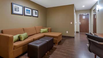 A living room with a sectional sofa, a leather ottoman, and a wooden coffee table. There are framed pictures on the wall and a door in the background.