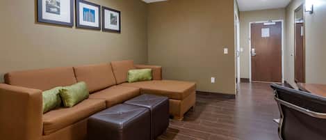 Una sala de estar con un sofá modular, un reposapiés de cuero y una mesa de centro de madera. Hay cuadros enmarcados en la pared y una puerta al fondo.