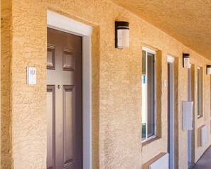 Un couloir avec des portes, des panneaux indiquant les numéros des pièces et des luminaires d’extérieur.