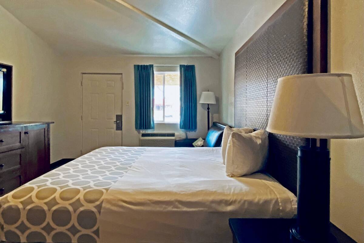 A hotel room with a bed, a desk, a lamp, a window with curtains, and a door.