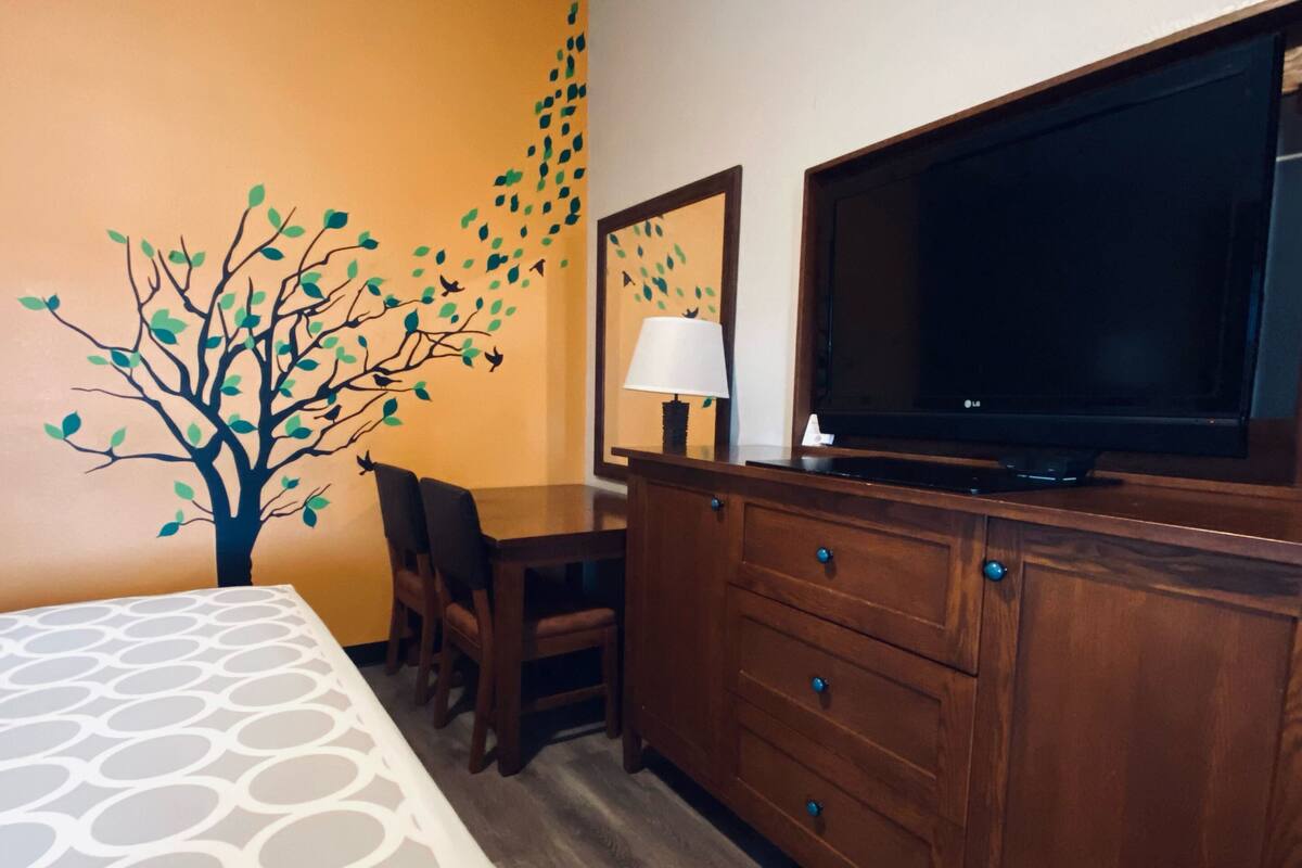 A hotel room with a bed, a wooden dresser, a television, and a wall with a tree decal.