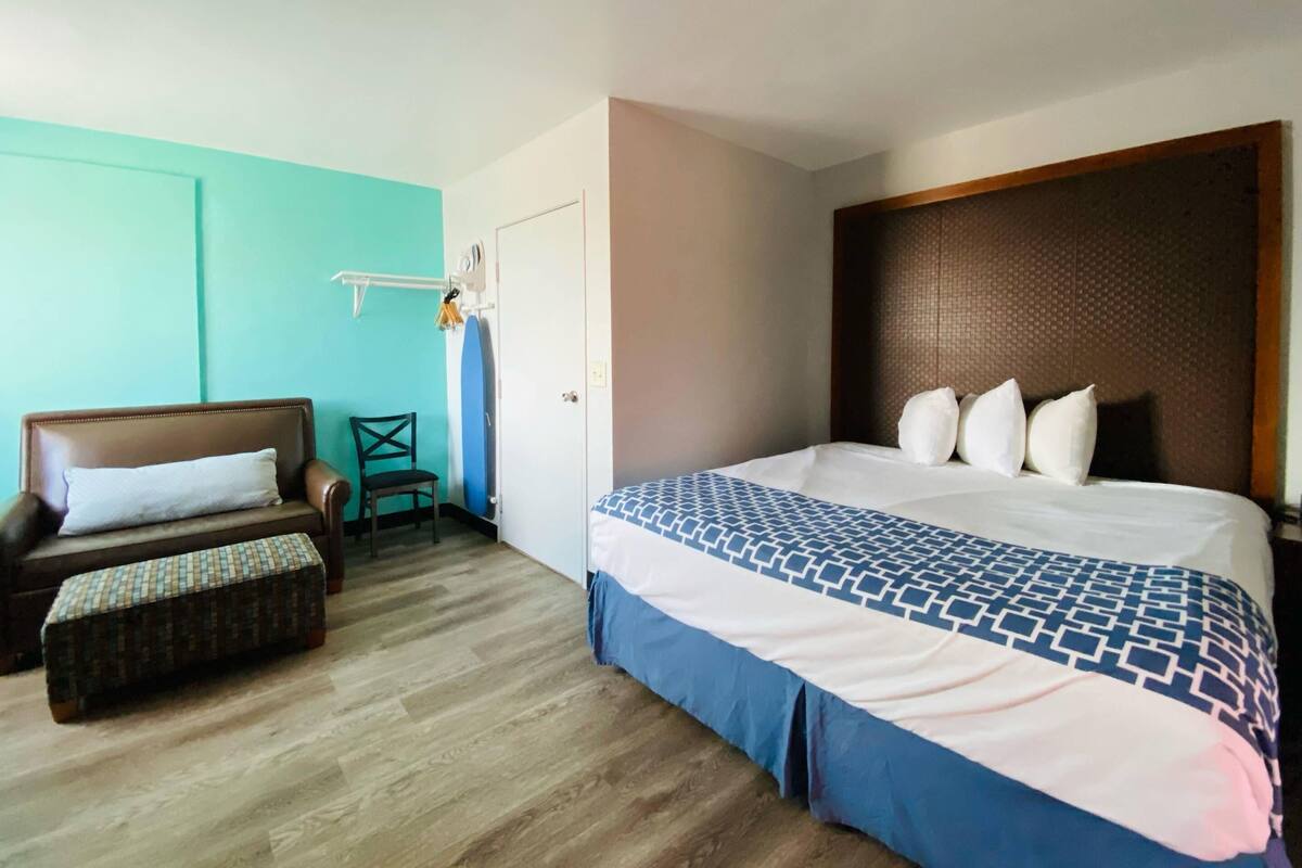 A hotel room with a bed, a chair, a small table, and a surfboard.