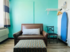 A room with a brown leather chair, a patterned ottoman, a black chair, a blue surfboard, and a wall-mounted air conditioning unit.