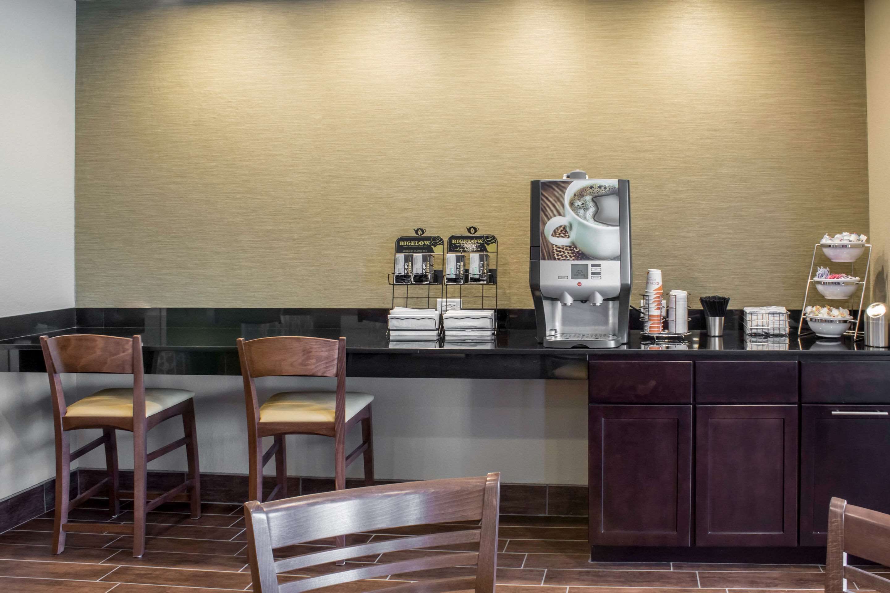 Una estación de café con dos máquinas de café, un dispensador de agua y un refrigerador pequeño. Hay dos sillas de madera y una encimera con varios artículos.