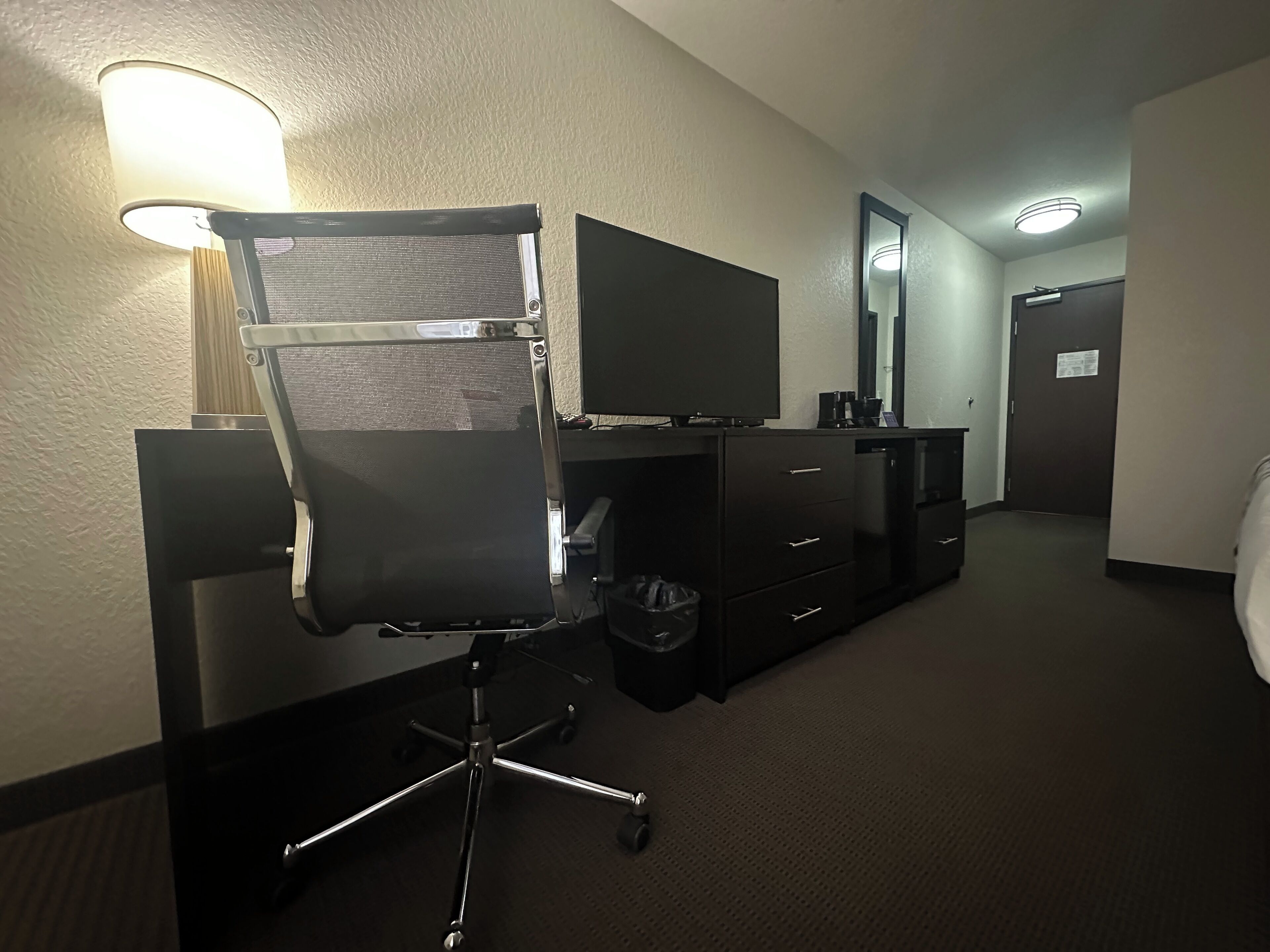 Habitación de hotel con una silla de oficina negra, un televisor de pantalla plana, un aparador, un cubo de basura y un espejo en la pared.