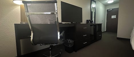 Habitación de hotel con una silla de oficina negra, un televisor de pantalla plana, un aparador, un cubo de basura y un espejo en la pared.