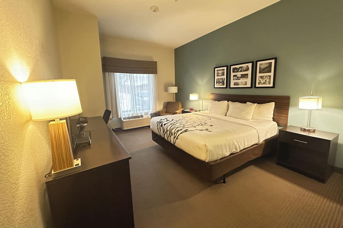 A hotel room with a large bed, bedside tables, a desk, and a chair. There are two windows with curtains, a wall-mounted lamp, and framed pictures on the wall.
