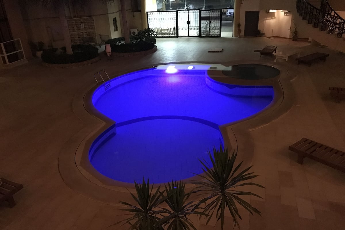 An indoor swimming pool with blue lighting, surrounded by lounge chairs and palm plants.