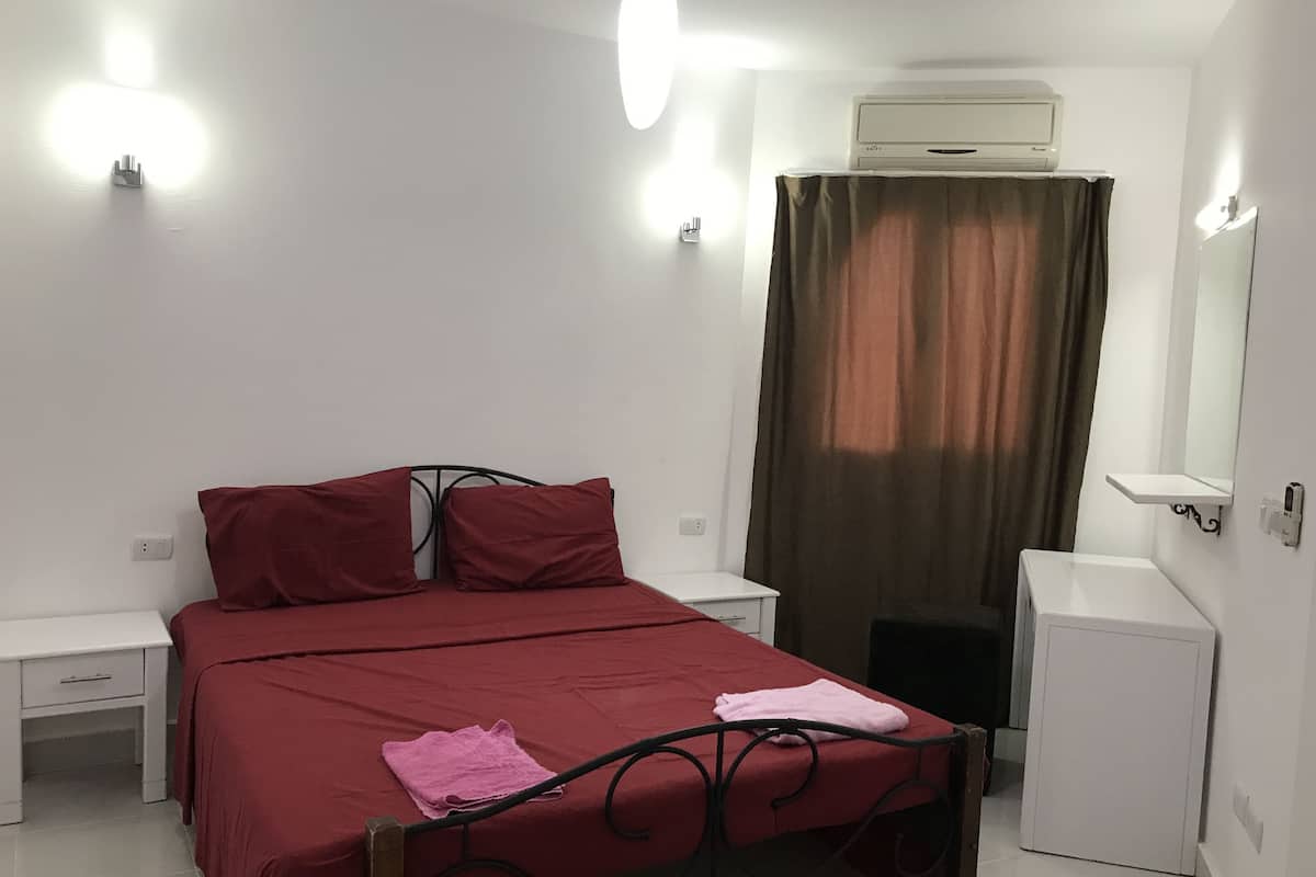 A bedroom with a bed, a bedside table, a small white cabinet, and an air conditioning unit.