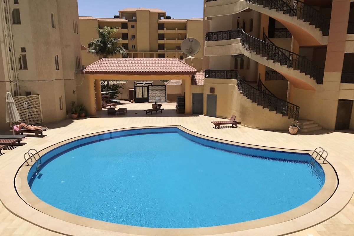 A swimming pool surrounded by lounge chairs and a covered seating area, with a staircase leading to an upper level.