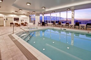 An indoor swimming pool with clear glass walls, lounge chairs, and a view of the cityscape. - Hampton Inn & Suites by Hilton Medicine Hat (Medicine Hat)