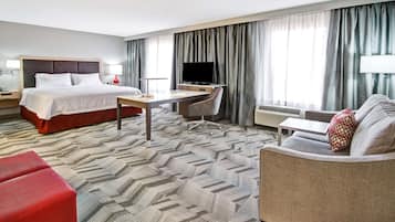 A hotel room with a bed, a desk, a chair, a sofa, a TV, and a window with curtains.