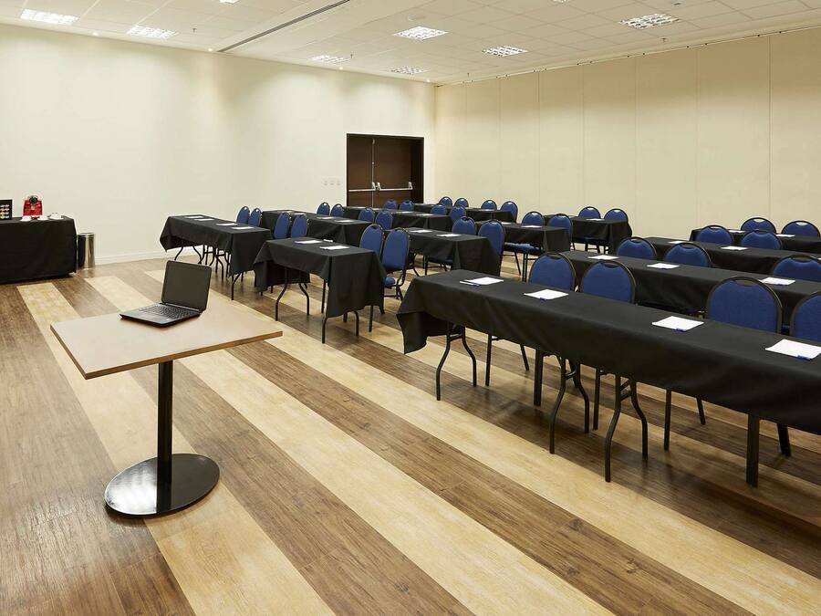 Un salón de conferencias con filas de mesas y sillas, un atril y una pequeña mesa con una computadora portátil y una planta.