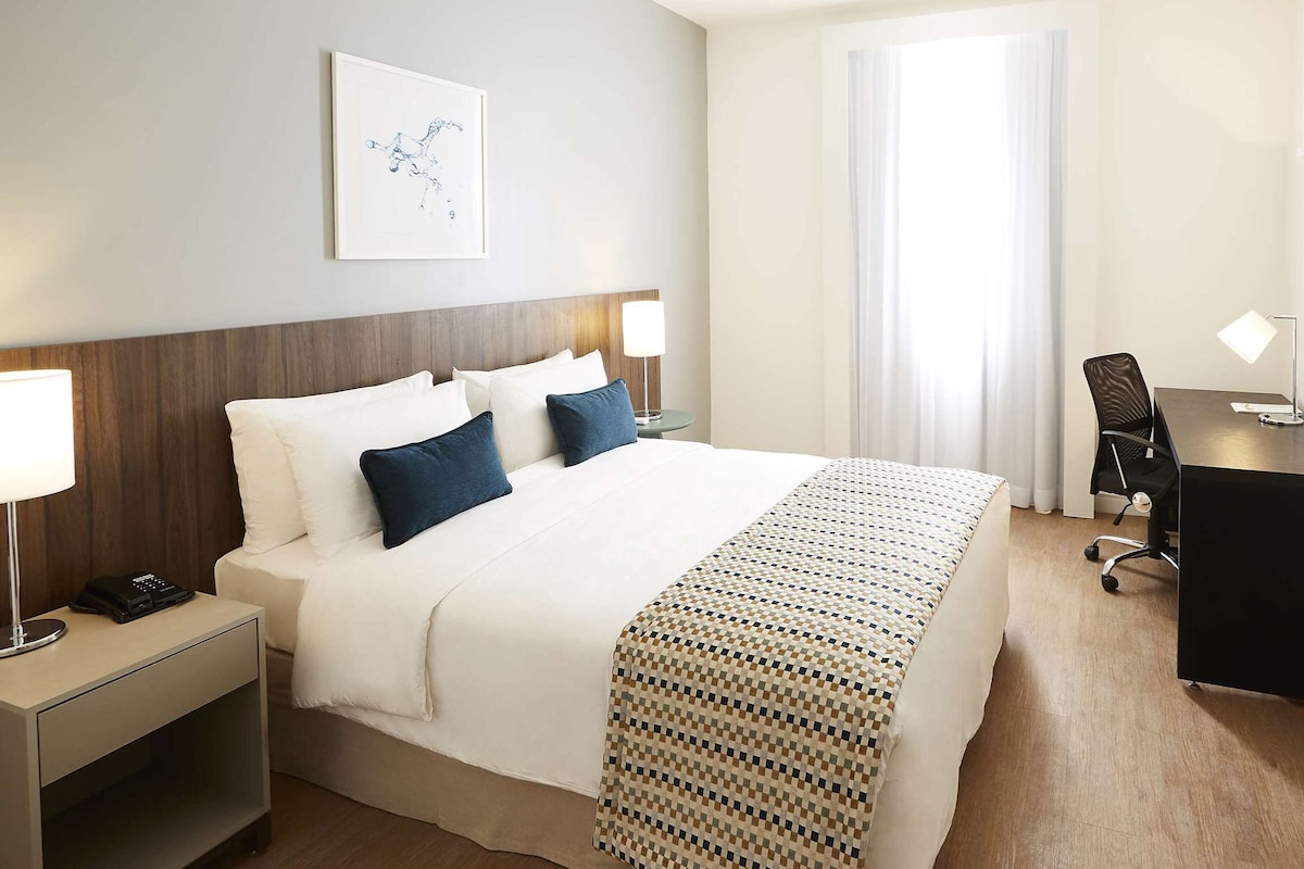 Habitación de hotel con cama, mesitas de noche, escritorio, silla, televisor y ventana con cortinas.