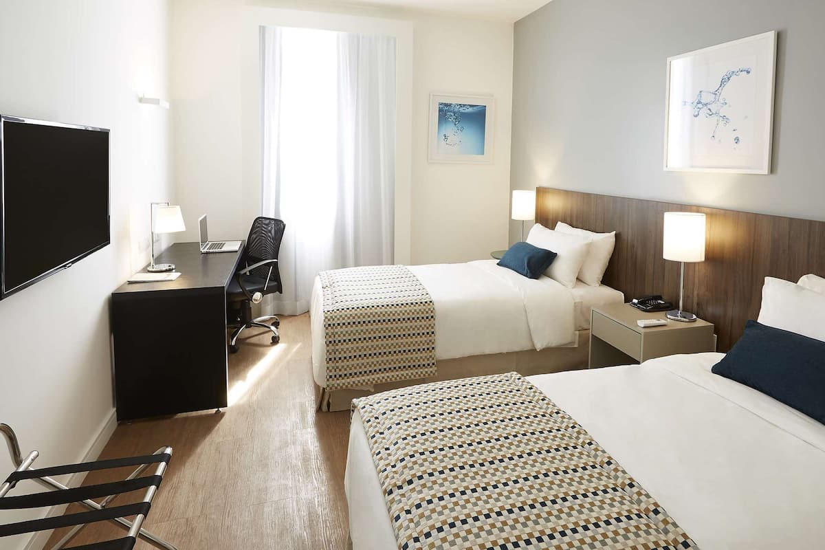 A hotel room with two beds, a desk, a chair, a TV, and a window with curtains.
